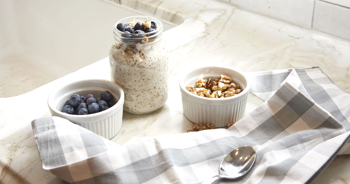 Small bowls of walnuts and blueberries sit next to a jar of overnight oats and a spoon resting on a tea towel.