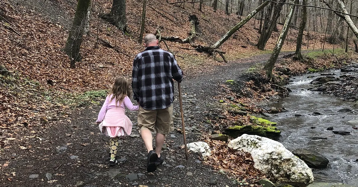 A father and daughter walk down a dirt trail next to a small creek. 