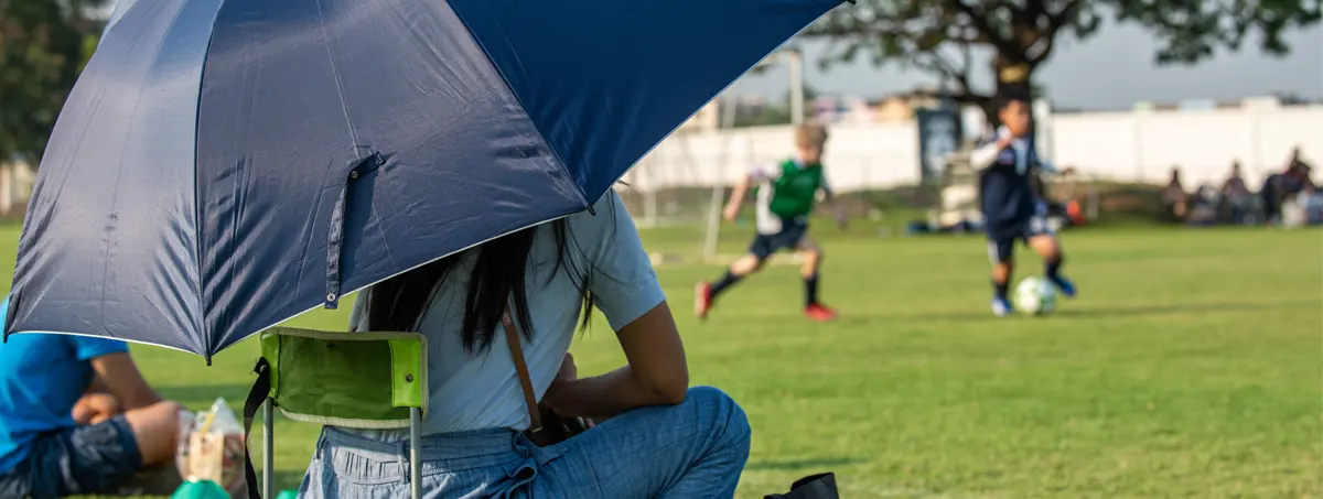 Woman at a kids soccer game sitting on chair and holding an umbrella.