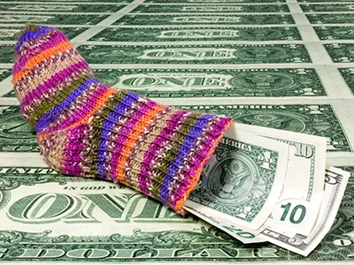 A colorfully striped hand knit sock laying on a sheet of money is filled with money of varying denominations. 