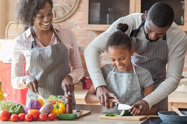 A family wearing aprons cooks together. A father helps his daughter slice a cucumber while the mom makes a salad. 
