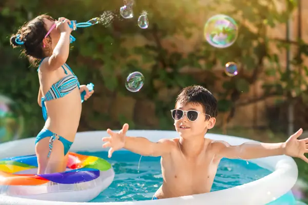 A little boy in a pool tries to catch bubbles blown by a little girl. 