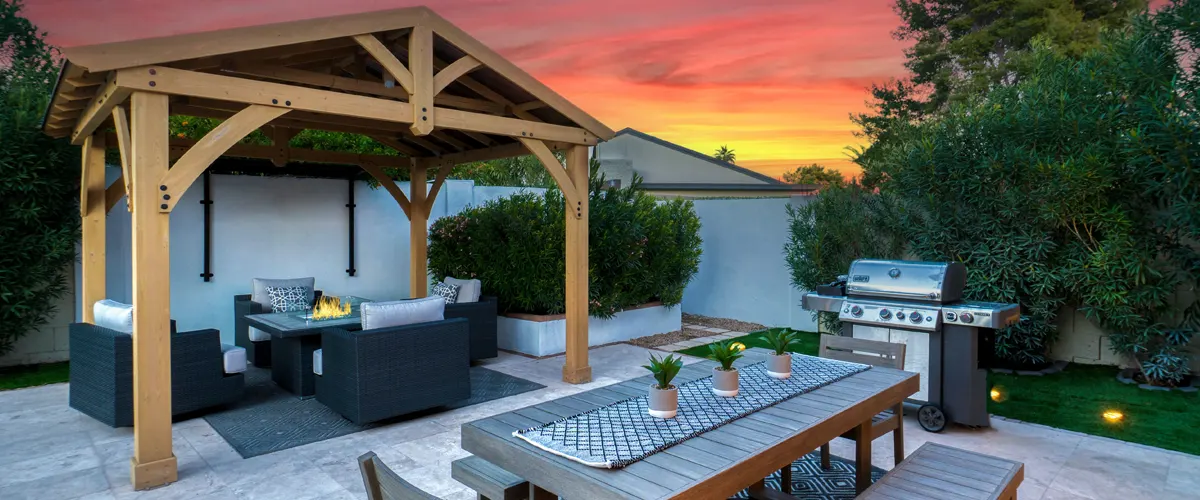 Outdoor patio scene at sunset with a grill, a pergola and patio furniture, and a dining table.