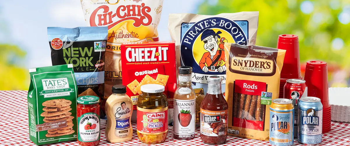 An outdoor table is filled with different snacks, beverages, grilling sauces, and condiments. 