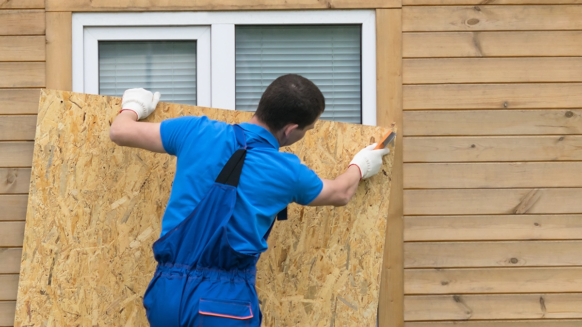 A man in blue overalls puts plywood over an exterior window before a storm.