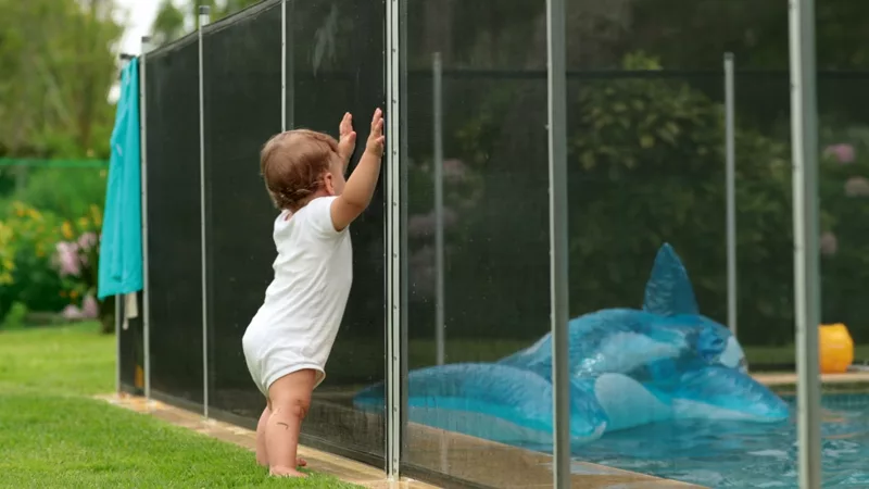 Baby leaning on safety fence surrounding a swimming pool