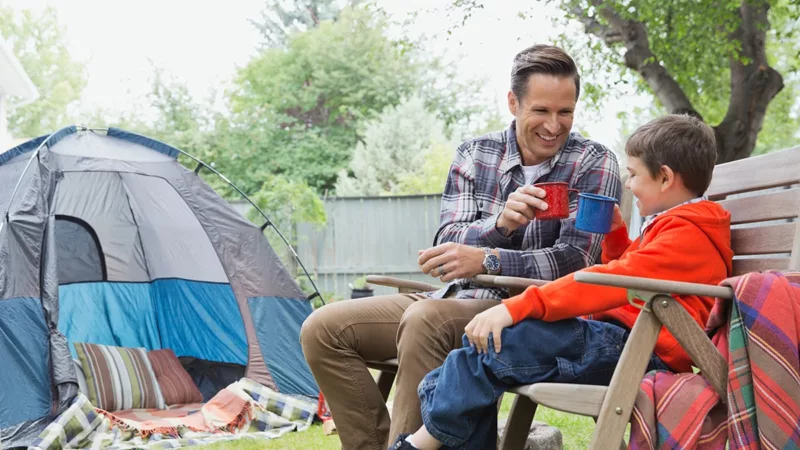 Father and son enjoy hot cocoa in backyard near a tent