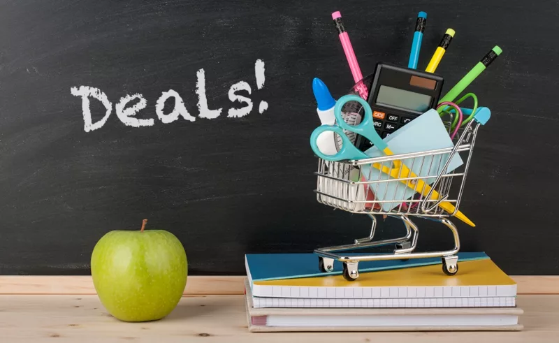 Next to a green apple is a small shopping cart filled with school supplies on top of a small stack of notebooks. A chalkboard reads "Deals!"
