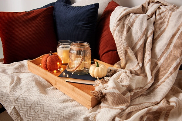 A sofa is covered with cream-colored blankets and rich jewel-toned throw pillows. A tray sits on top with candles, a book, and fairy lights.
