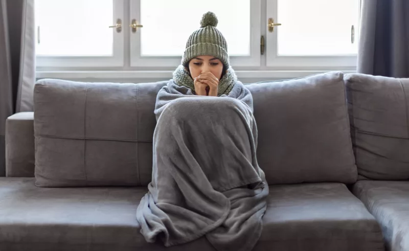 A chilly person wrapped in a blanket on a couch blowing into their hands