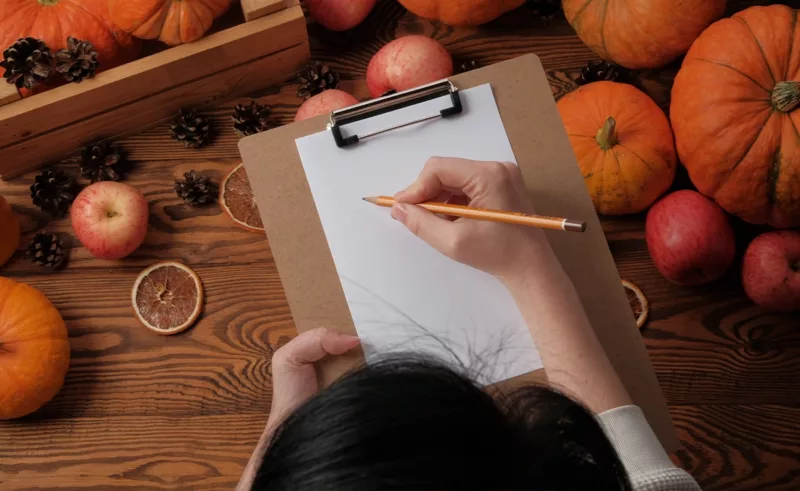 A woman starts a list while sitting at a table filled with pumpkins and apples.