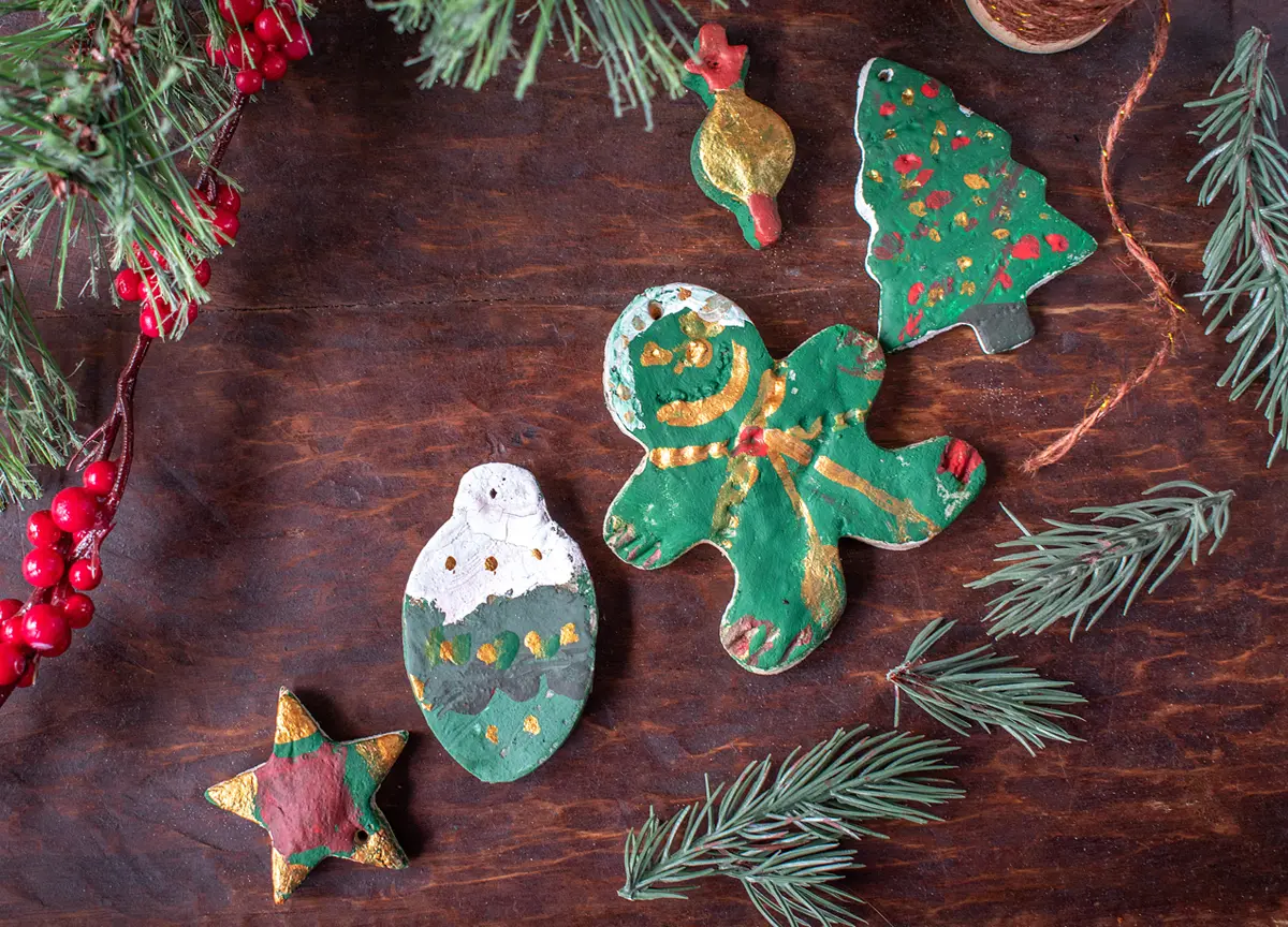 Handpainted salt dough ornaments on a wooden table surrounded by pine greens and holly berries.