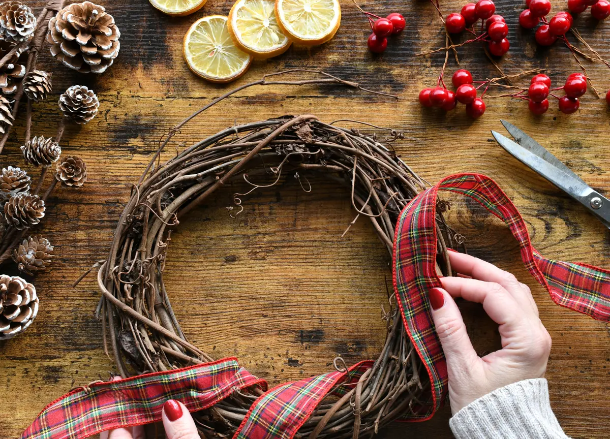 A woman decorates a grapevine wreath with a red, green, and gold plaid ribbon.