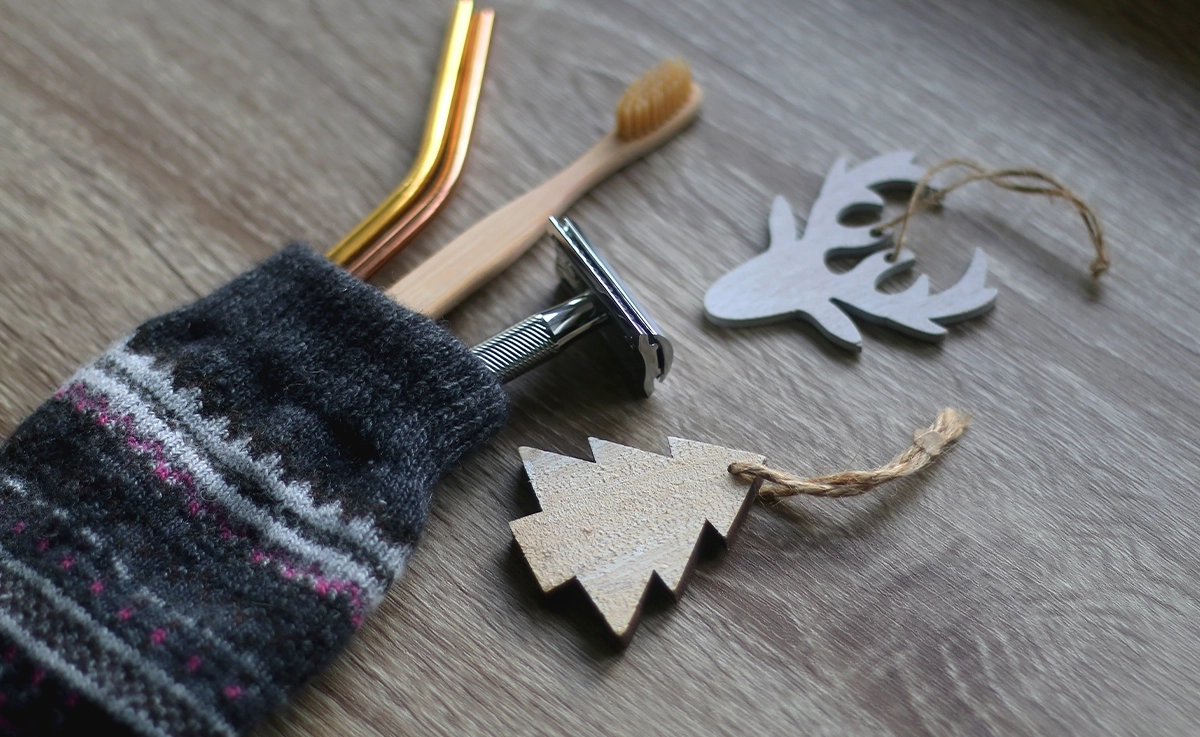 A Christmas stocking filled with reusable straws, a wooden toothbrush, and a reusable metal razor lay next to wooden Christmas ornaments.