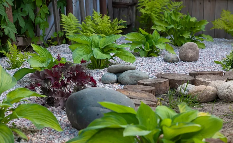 A gravel path is lined with stones, plants, and wooden cylinders.