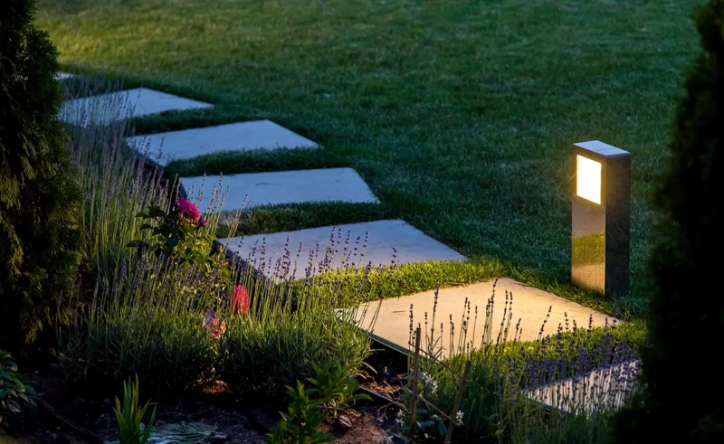 A garden path made of pavers is illuminated by a light at dusk.