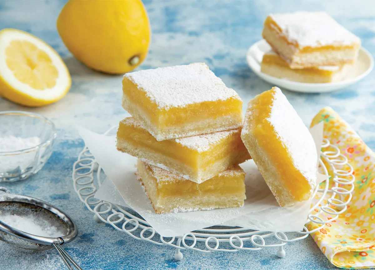 A plate of lemon bars sits on a table surrounded by powdered sugar and lemons.