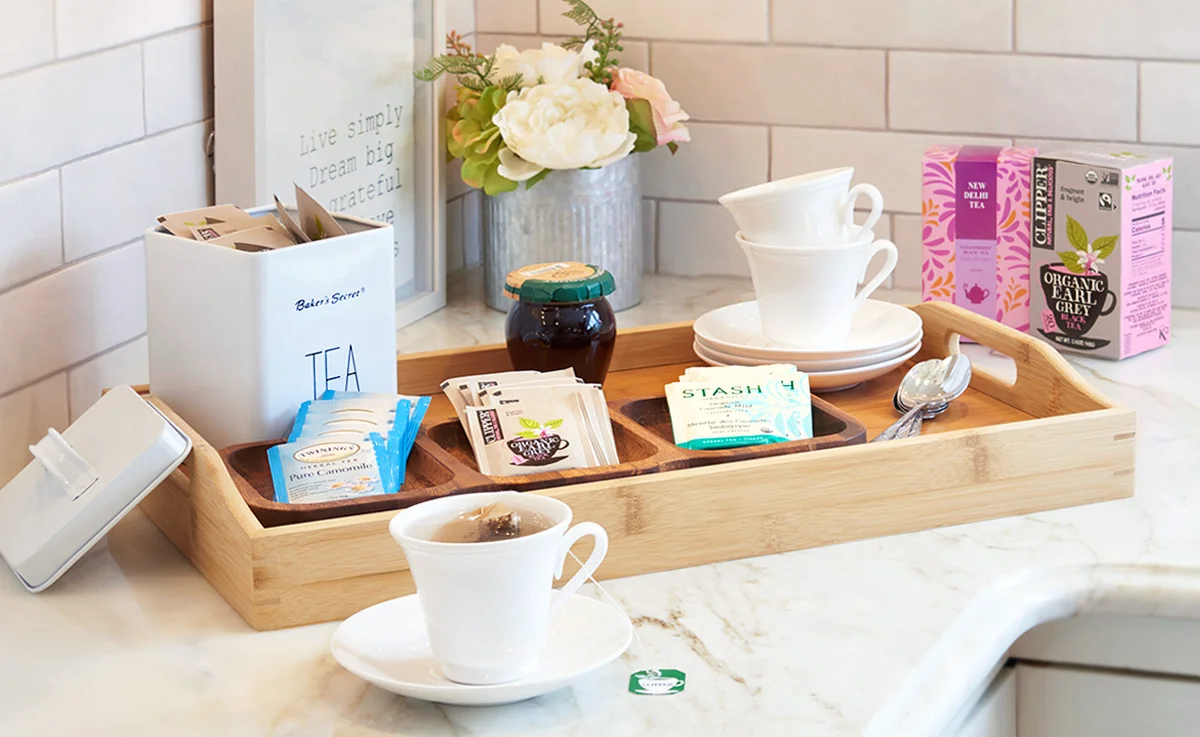 A wooden tray filled with tea cups, tea bags, and spoons sits on a kitchen countertop