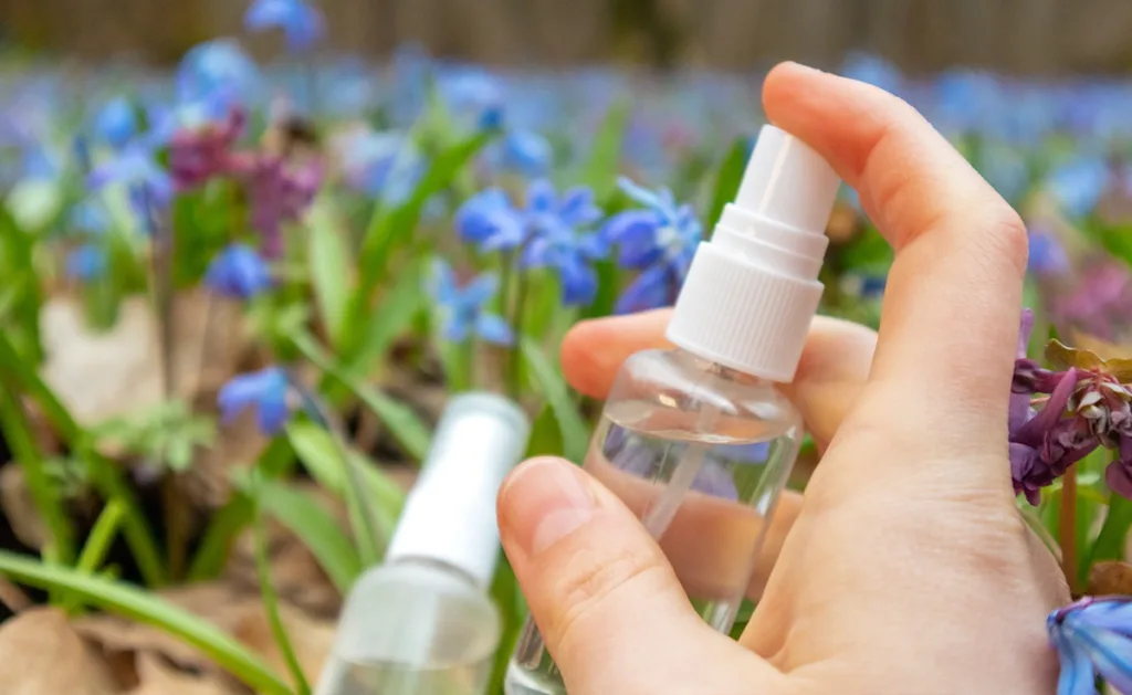 A hand holding a clear spray bottle, with plants in the background.