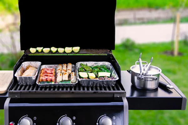 An opened gas grill with vegetables, meat, and sausages in aluminum barbeque trays.