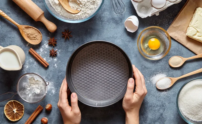 A top-down view of a person holding a baking pan surrounded by baking tools and common baking ingredients.