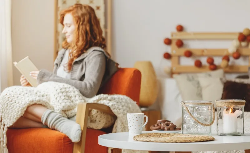 A person sitting on an armchair reading a book under a cozy blanket next to tea and candles.