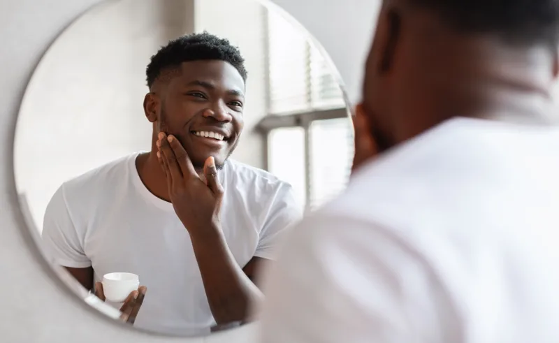 A person looks in a mirror while applying lotion to their face.