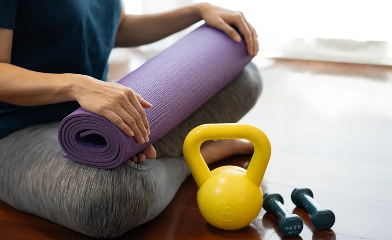 A person holds a yoga mat, sitting next to a kettlebell and some hand weights.