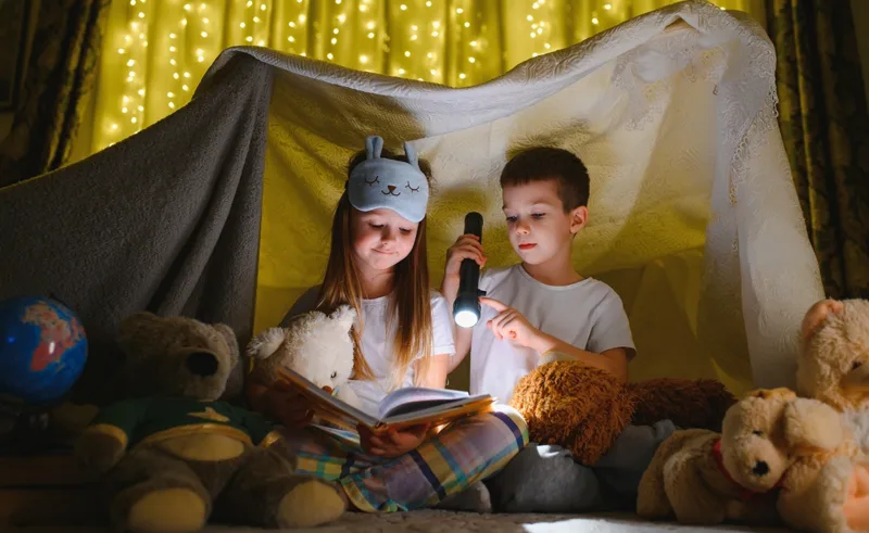 Two kids reading a book by flashlight in a cozy blanket fort.