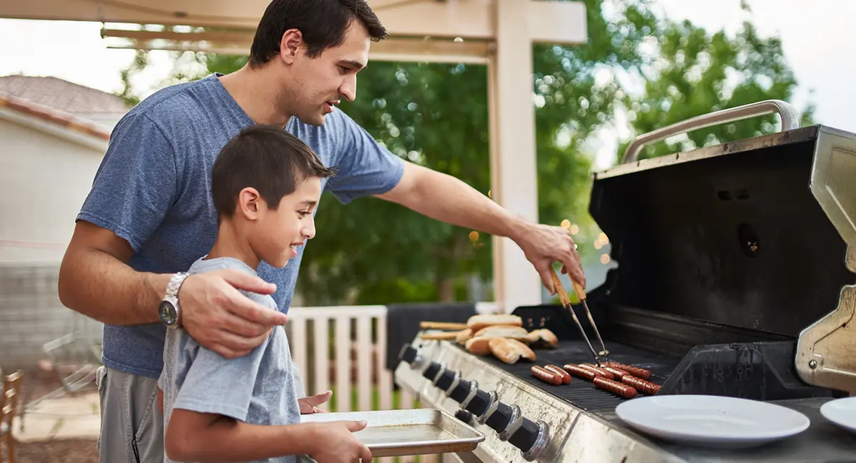 A father teaching his son how to grill hot dogs and bonding over cooking on the grill.